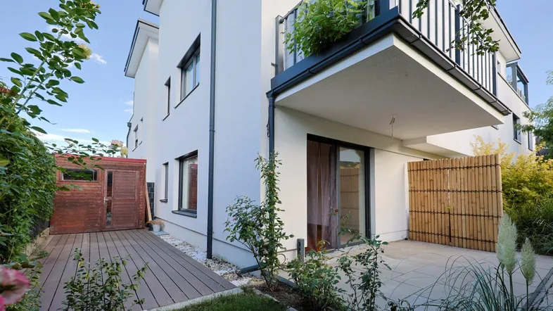 Moderne Hausfassade mit weißem Putz, angrenzender Terrasse und Holzdeck im Gartenbereich.