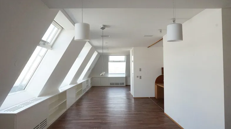 Heller Dachbodenraum mit schrägen Wänden, mehreren Dachfenstern und dunklem Holzboden.