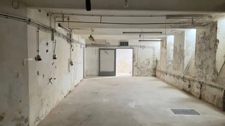Großer, leerer Kellerraum mit Betonboden und sichtbaren Leitungen, Wände zeigen Abnutzungsspuren.