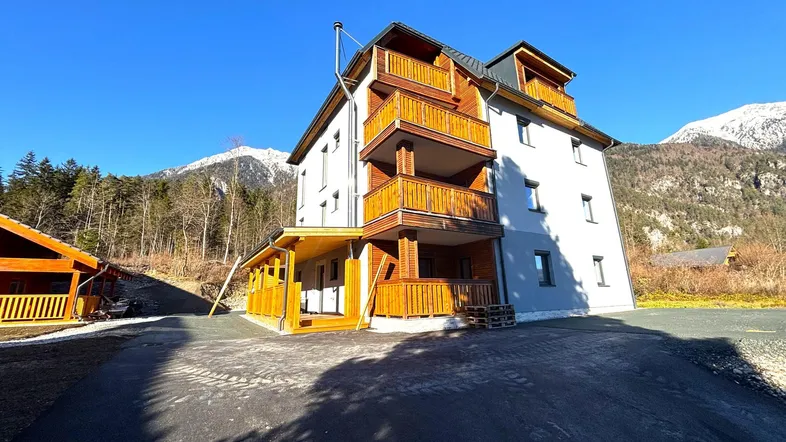Modernes Mehrfamilienhaus mit Holzbalkonen und Bergblick unter strahlend blauem Himmel.