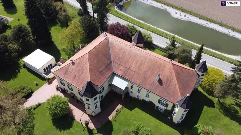 Luftaufnahme eines weitläufigen Anwesens mit historischem Gebäude, umgeben von viel Grün und einem Teich.