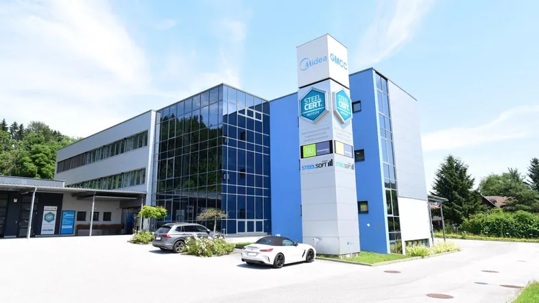 Modernes Bürogebäude mit Glasfassade und blauen Akzenten, umgeben von Grünflächen und Parkplätzen.