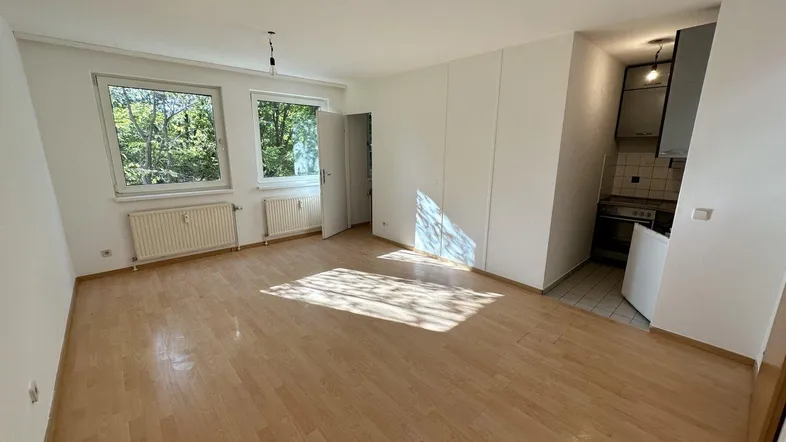 Helles Zimmer mit Parkettboden und Blick ins Grüne, offen verbunden mit einer kleinen Küchenzeile.