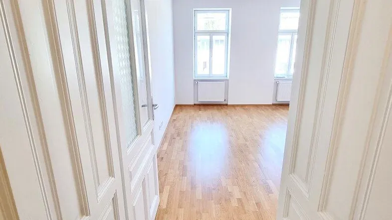 Heller Wohnraum mit Parkettboden und hohen Fenstern, gesehen durch eine offene Tür.