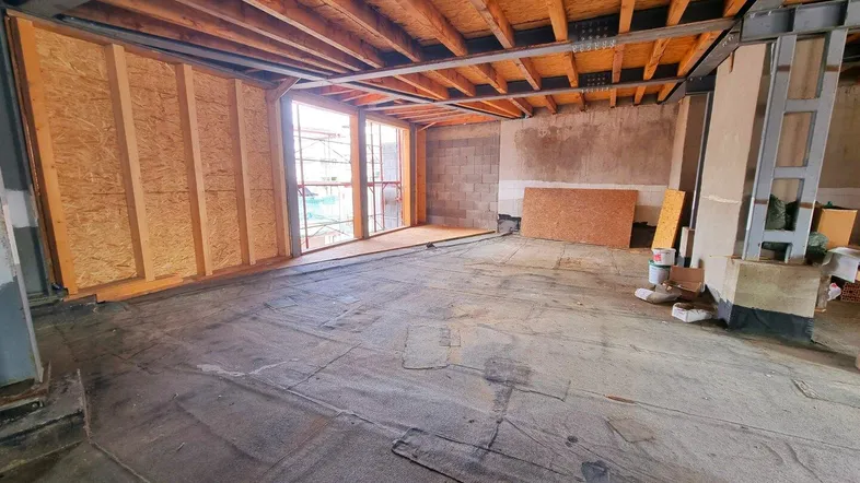 Großer Innenraum im Rohbau mit sichtbaren Holz- und Metallkonstruktionen sowie großen Fensteröffnungen.