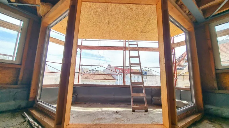 Rohbau eines Dachgeschossraumes mit großen Fensteröffnungen und sichtbaren Holzbalken während der Bauphase.
