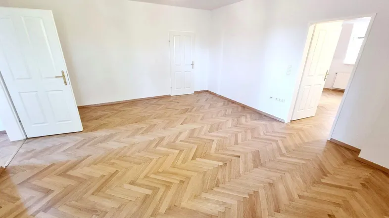 Heller, leerer Raum mit klassischem Fischgrätparkett und weißen Wänden.