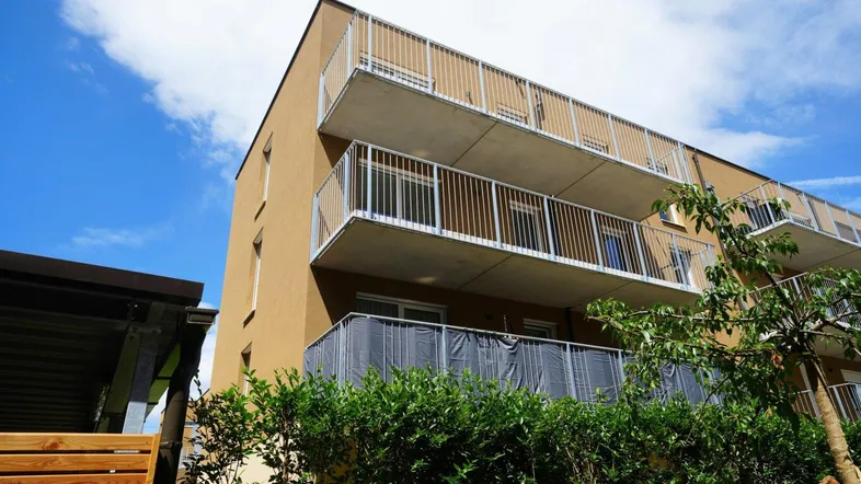 Modernes Mehrfamilienhaus mit mehreren Balkonen unter blauem Himmel, umgeben von grünen Büschen.