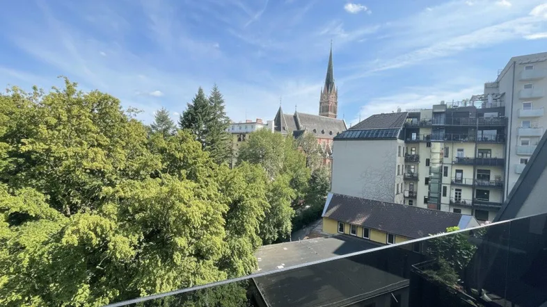 Blick von einem Balkon auf eine Stadtlandschaft mit üppigem Grün und einer historischen Kirche unter blauem Himmel.