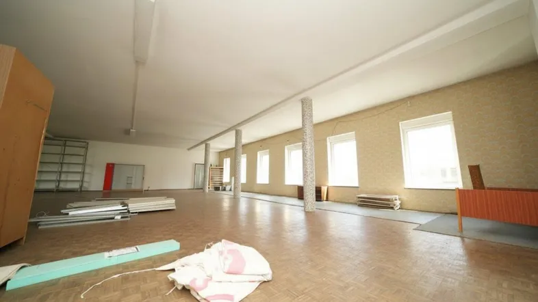 Großer, leerstehender Gewerberaum mit Parkettboden, vielen Fenstern und Regalen an der Wand.
