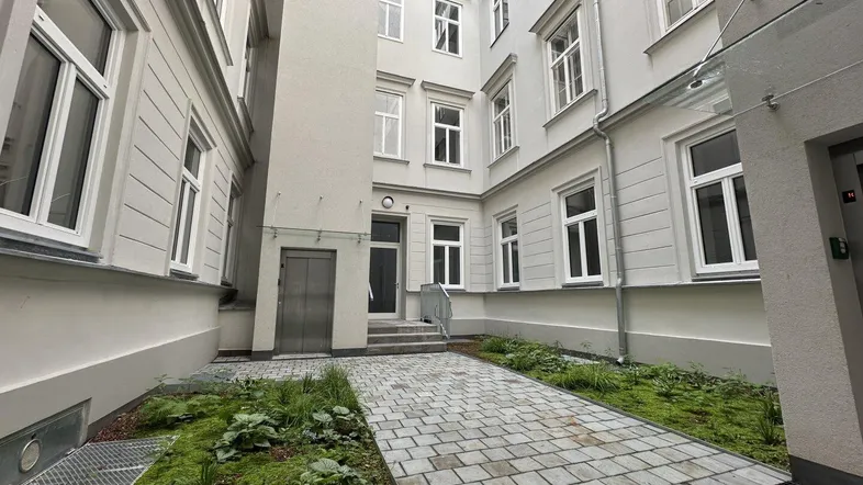 Heller Innenhof eines Wohngebäudes mit gepflastertem Weg, Grünflächen und Zugang zum Eingang.