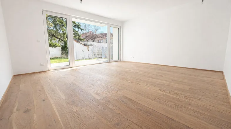 Helles, leeres Zimmer mit großen Fenstern, Holzboden und Zugang zu einem Gartenbereich.