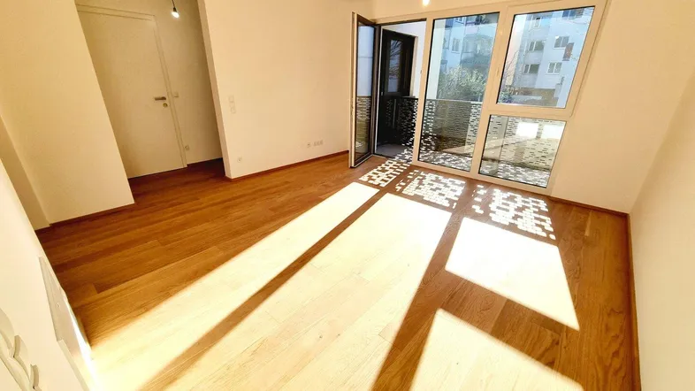 Helles, leeres Zimmer mit Holzboden und Zugang zu einem Balkon, durchflutet von Sonnenlicht.