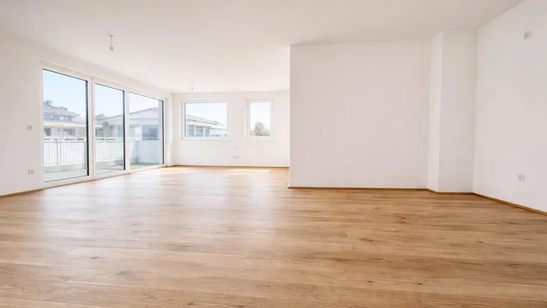 Heller, leerer Wohnbereich mit großen Fenstern, Holzboden und Zugang zu einem Balkon.