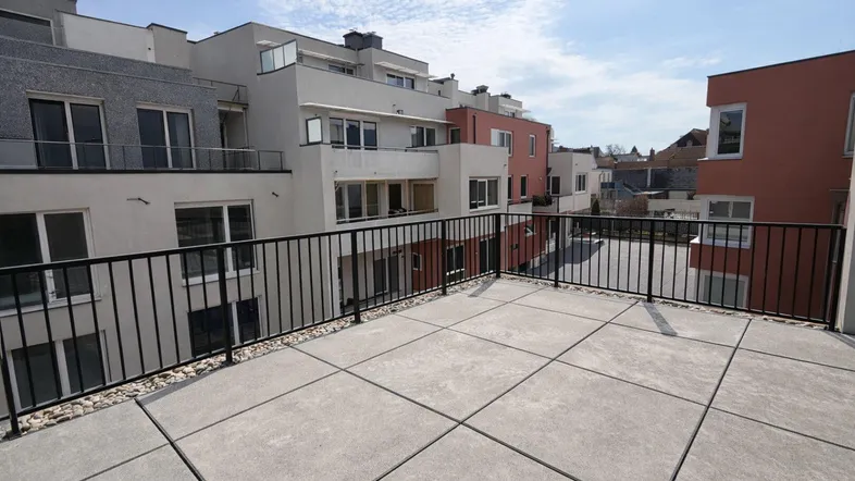 Geräumige Dachterrasse mit Fliesenboden und Blick auf moderne Wohngebäude unter blauem Himmel.