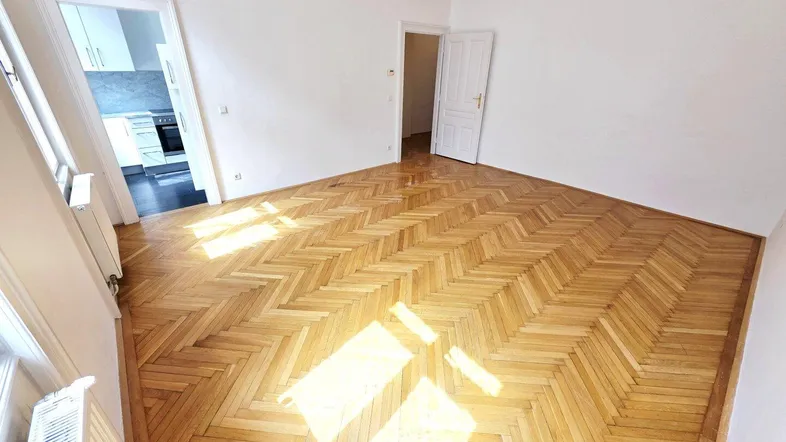 Helles Zimmer mit Fischgrätparkettboden und Zugang zu einem weiteren Raum.
