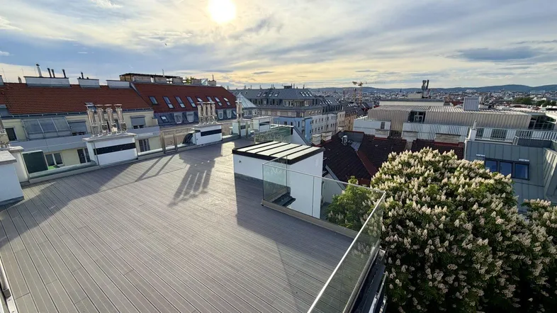 Große Dachterrasse mit modernem Bodenbelag, Glasgeländer und weitem Stadtblick unter sonnigem Himmel.