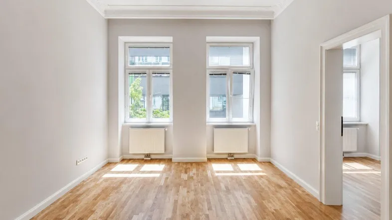 Helles Zimmer mit Parkettboden, hohen Decken und zwei großen Fenstern, ideal als Wohn- oder Schlafzimmer.