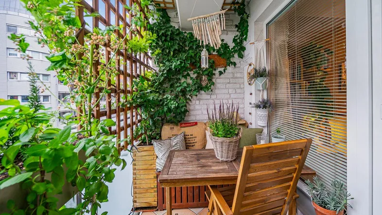 Sonniger Balkon mit Holztisch und Stühlen, umgeben von üppigen Pflanzen und einer Ziegelwand.