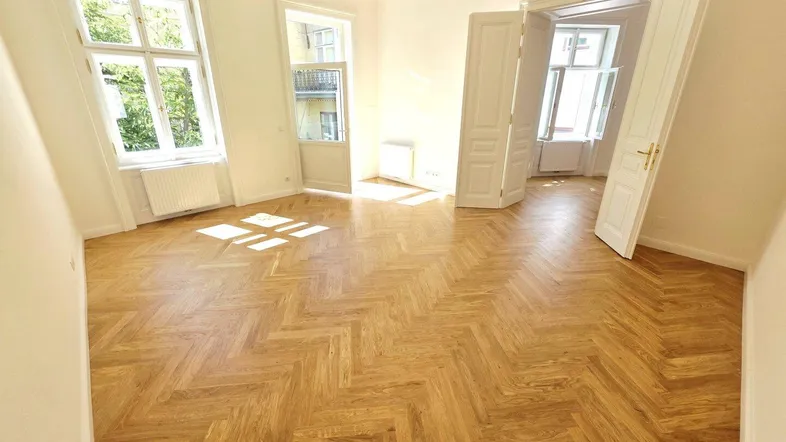Helles Zimmer mit Fischgrätparkett und hohen Fenstern, das viel Tageslicht hereinlässt.