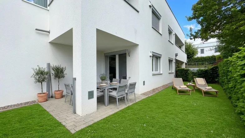 Möblierte Terrasse mit Rasenfläche und Liegestühlen vor dem modernen Gebäude.
