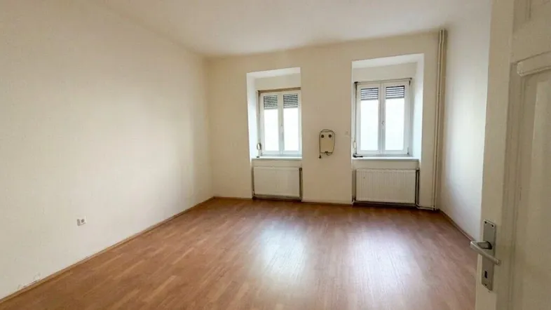 Helles, leeres Zimmer mit Laminatboden und zwei Fenstern, bereit zur individuellen Gestaltung.