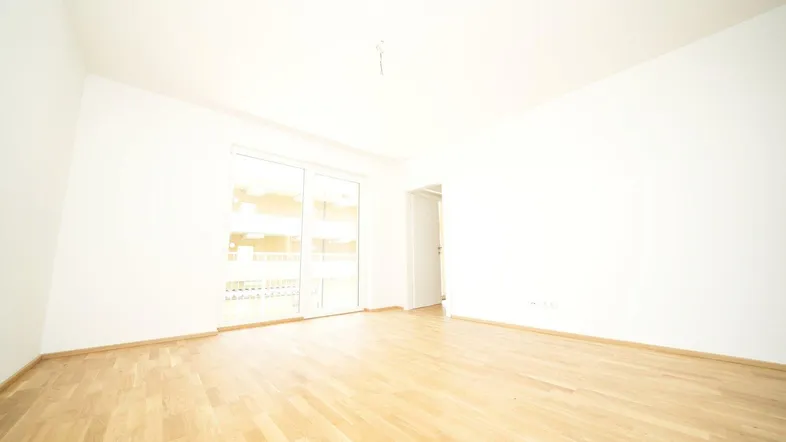 Helles, leeres Zimmer mit Parkettboden und Zugang zu einem Balkon oder einer Terrasse.