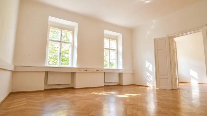 Heller, leerer Raum mit Fischgrätparkett, zwei Fenstern und weißen Wänden, gut beleuchtet.
