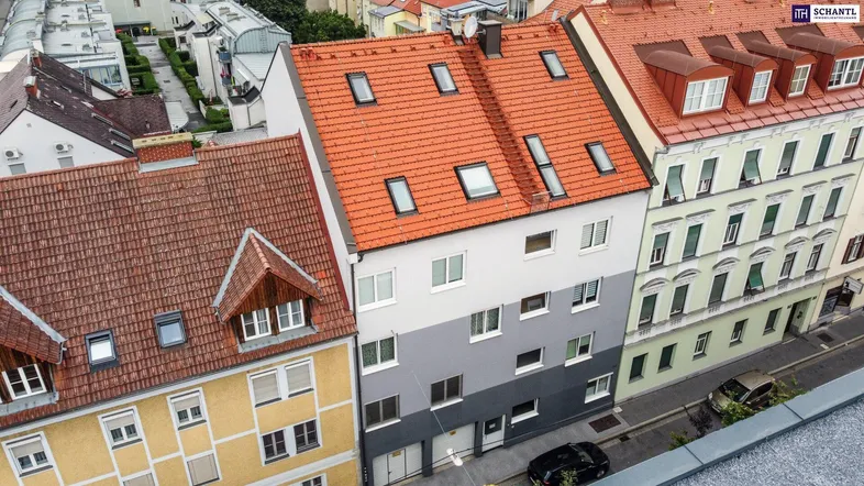 Luftaufnahme eines Mehrfamilienhauses mit rotem Ziegeldach und Dachfenstern in städtischer Umgebung.