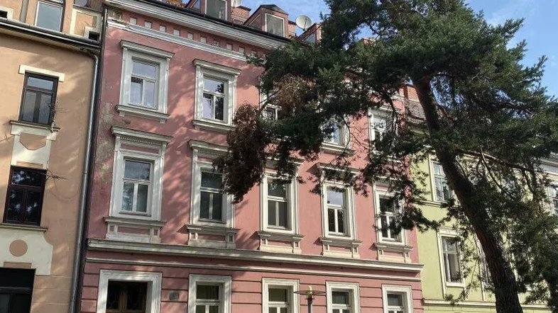 Historisches Gebäude mit rosafarbener Fassade, grüner Hecke und einem Baum davor.