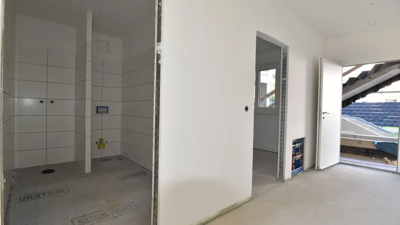 Unfertiges Badezimmer mit weißen Fliesen und Rohinstallationen, daneben ein weiterer Raum im Rohbau.