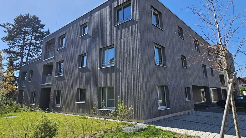 Modernes Mehrfamilienhaus mit Holzfassade und gepflegter Grünfläche unter blauem Himmel.
