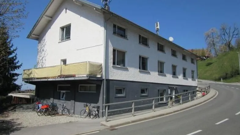 Weißes Mehrfamilienhaus mit dunklem Sockel und Balkon an einer Straße gelegen.