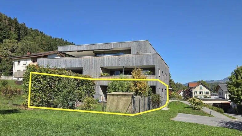 Modernes Mehrfamilienhaus mit Holzfassade und umzäuntem Gartenbereich in grüner Umgebung.