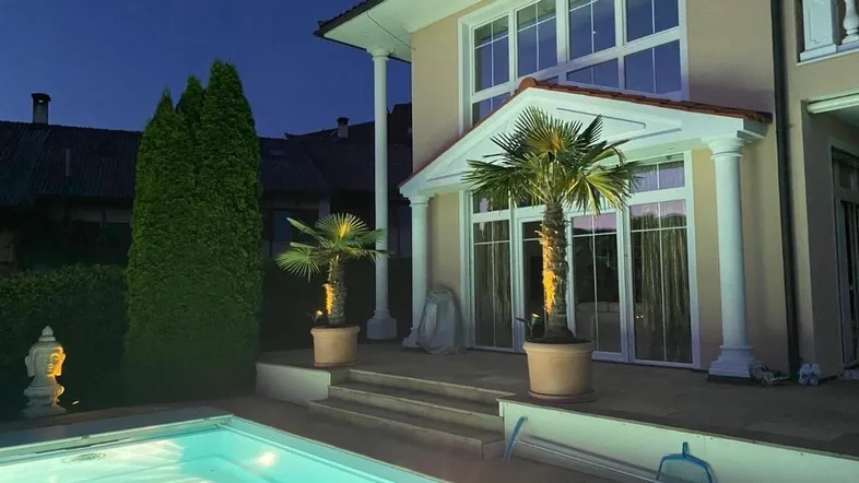 Beleuchteter Poolbereich vor einer eleganten Villa bei Nacht mit Palmen und großen Fenstern.