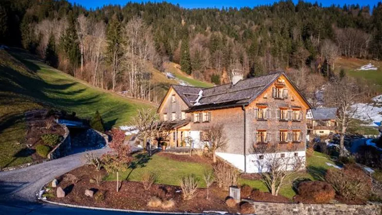 Traditionelles Holzhaus in alpiner Landschaft mit umliegenden Hügeln und Wald unter blauem Himmel.