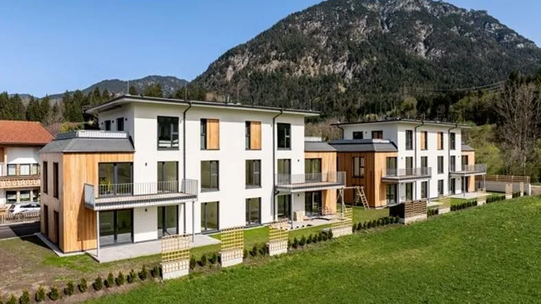 Moderne Mehrfamilienhäuser mit weißer Fassade und Holzverkleidung, umgeben von grünen Wiesen und Bergen.