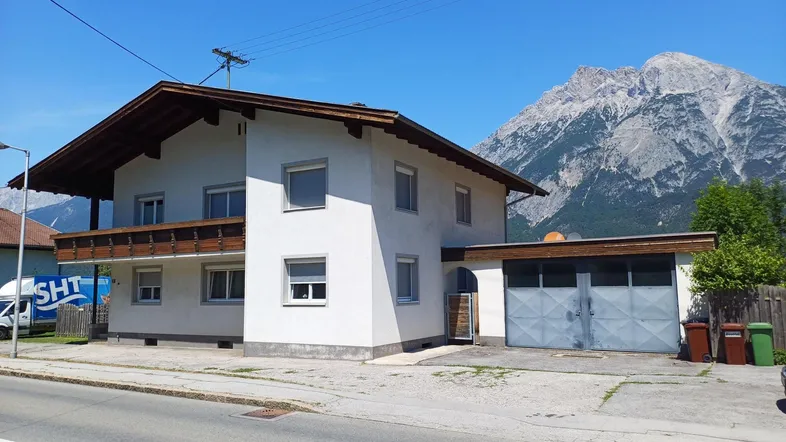 Wohnhausfassade mit Balkon und traditionellem Holzdach vor einer beeindruckenden Bergkulisse.