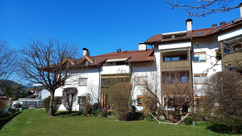 Mehrfamilienhaus mit weißer Fassade, braunem Dach und gepflegtem Vorgarten unter blauem Himmel.