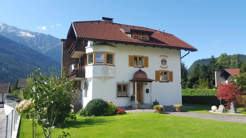 Traditionelles Haus im alpinen Stil mit Bergblick und gepflegtem Vorgarten unter blauem Himmel.