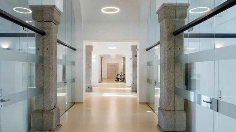 Langer, heller Flur mit hohen Gewölbedecken, Steinsäulen und modernen Glaswänden.
