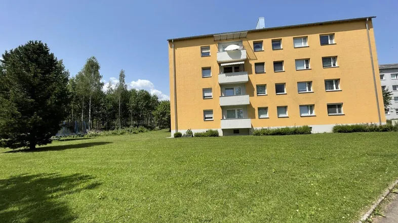 Gelbes Mehrfamilienhaus mit Balkonen und weitläufiger Grünfläche unter blauem Himmel.