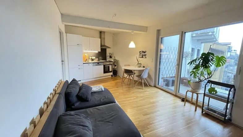 Heller offener Wohn- und Essbereich mit integrierter Küche, Holzboden und Zugang zum Balkon.