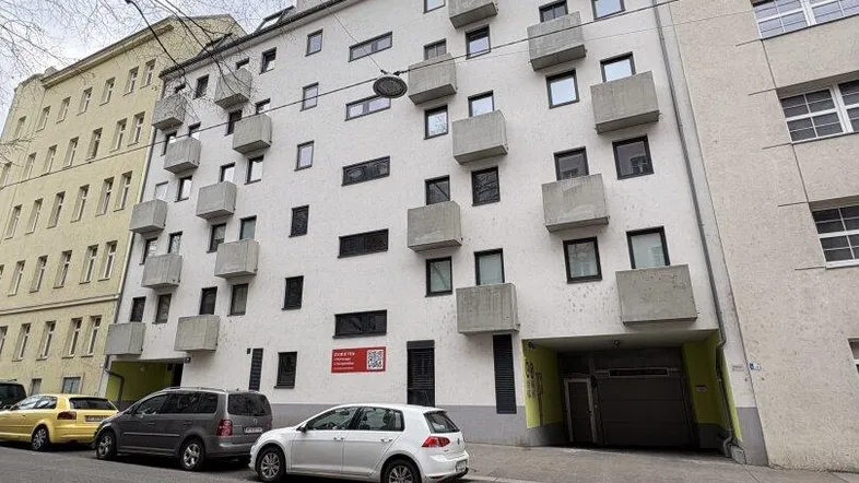 Mehrstöckiges Wohngebäude mit zahlreichen kleinen Balkonen und parkenden Autos auf der Straße.