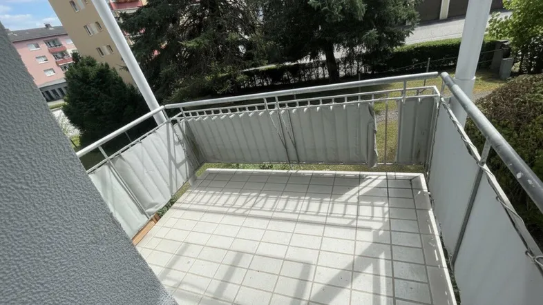 Heller Balkon mit Fliesenboden und seitlichen Sichtschutzelementen, umgeben von Grün.