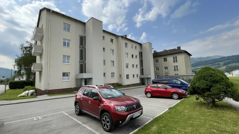 Mehrfamilienhaus mit beige gestrichener Fassade und Parkplätzen davor unter bewölktem Himmel.