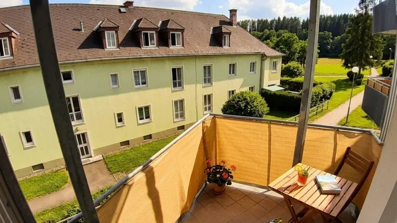 Sonniger Balkon mit Gartenmöbeln und Blick auf die grüne Umgebung und ein weiteres Gebäude.