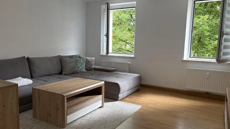 Helles Wohnzimmer mit modernem Sofa, Holzboden und Blick ins Grüne durch offene Fenster.