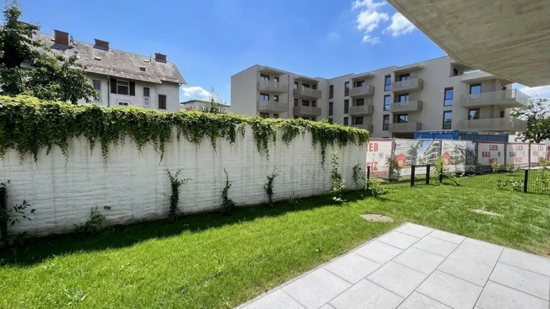 Gepflegter Gartenbereich mit Rasenfläche und gepflasterter Terrasse, umgeben von einer begrünten Mauer.