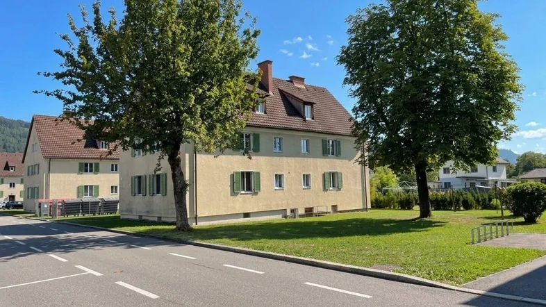 Mehrfamilienhaus mit heller Fassade, grünen Fensterläden und gepflegter Umgebung unter blauem Himmel.